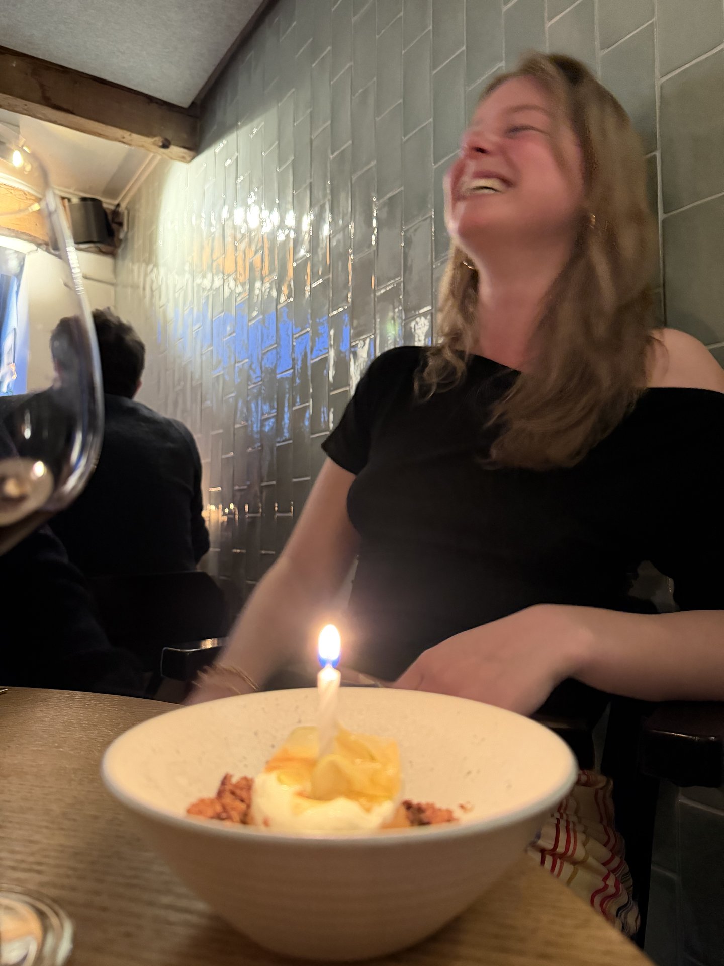 A woman is captured mid-laugh, celebrating with a dessert topped with a lit candle in a cozy restaurant at Govert Flinckstraat, Oude Pijp, Amsterdam